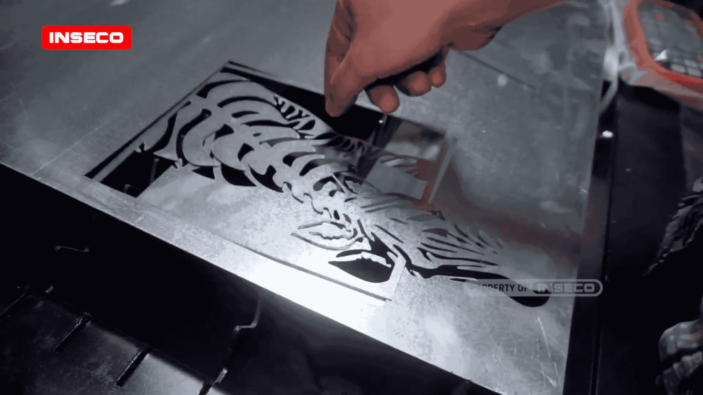 Tampilan dekat tangan yang sedang mengambil desain logam yang telah dipotong dari meja pemotongan laser, menyoroti detail rumit dari desain yang telah selesai.